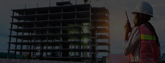 Female construction supervisor using a walkie-talkie in front of a high-rise building under construction at sunset.