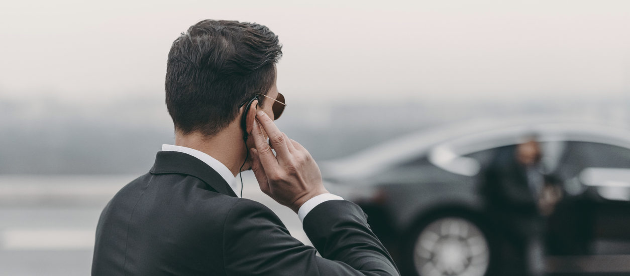 Professional security guard in suit speaking into earpiece while standing beside a black vehicle